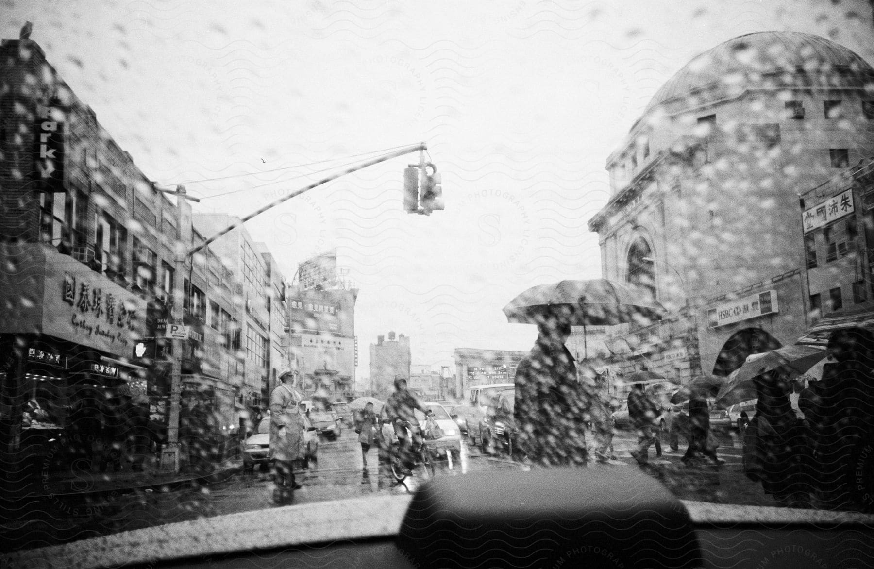 A black and white view of a city street with people walking holding umbrellas and buildings lined on either side with a fourway traffic light above the road