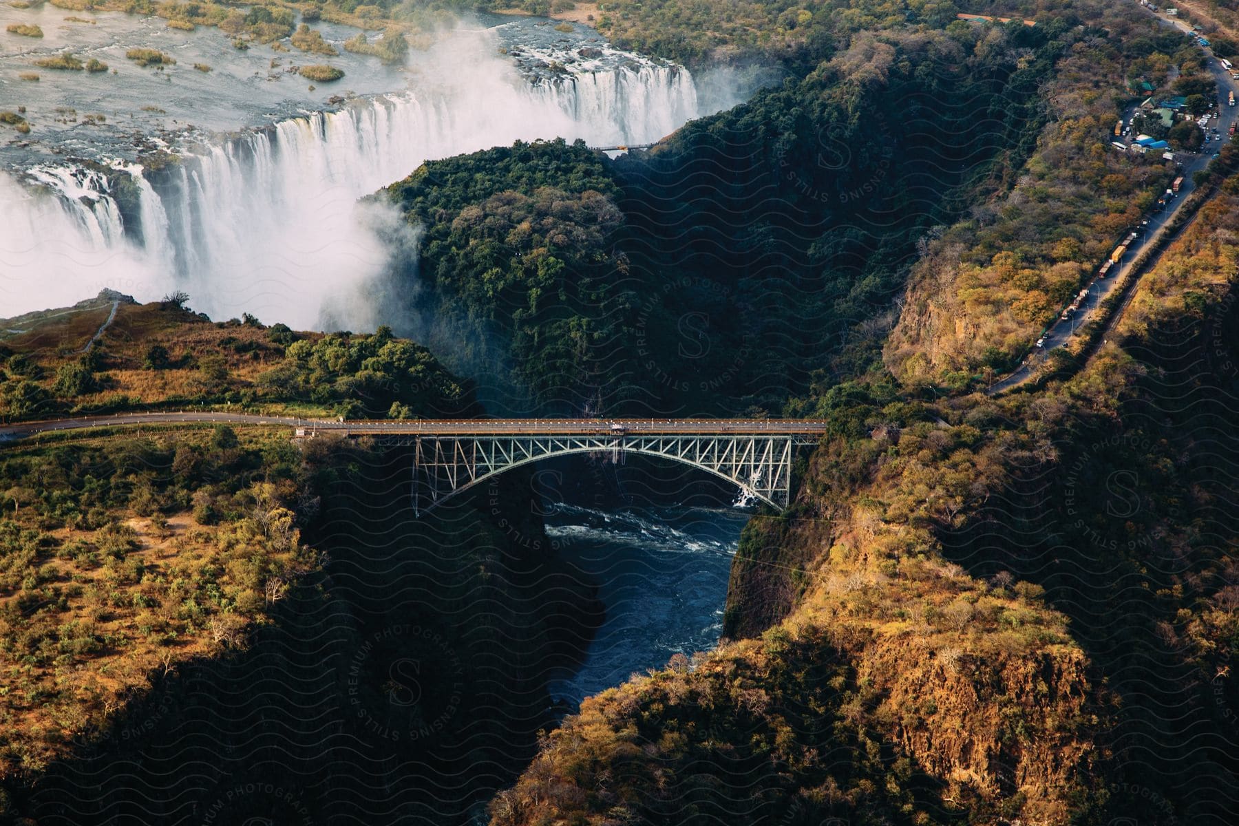 An automobile crosses a bridge over a deep ravine with a river below as waterfalls flow over a cliff