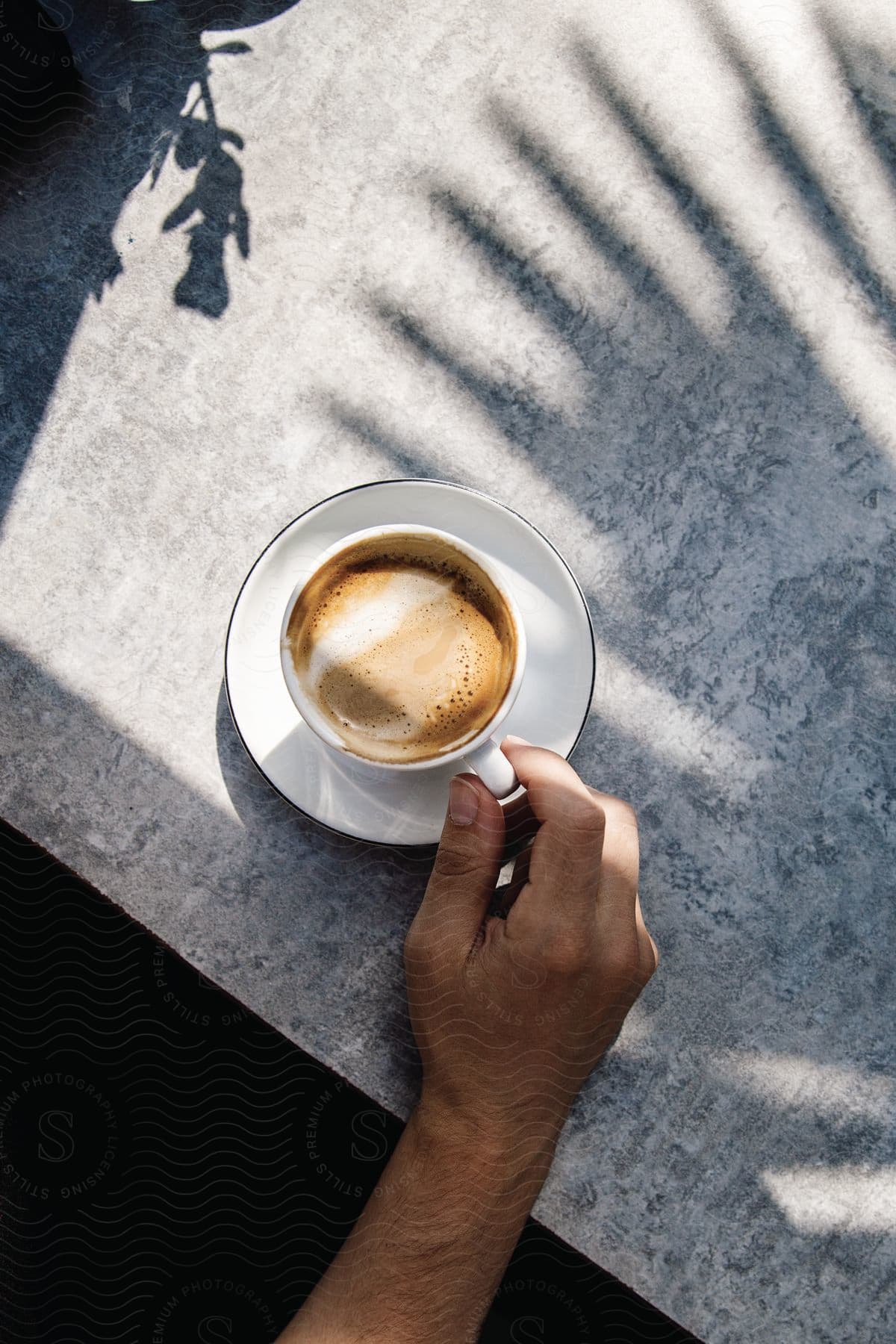 A mans hand holding a white coffee cup on a grey table on a sunny day