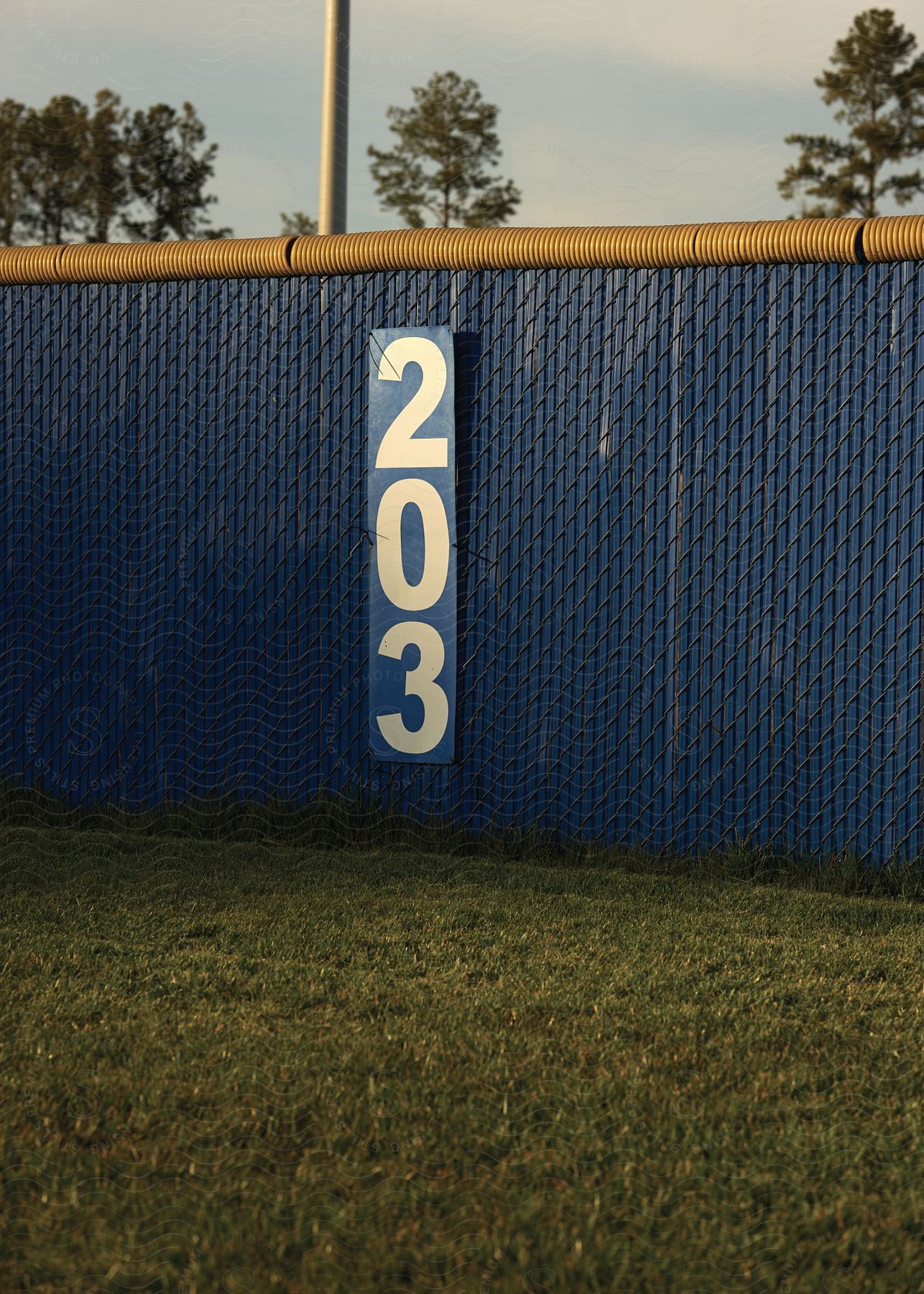 A blue chain link fence with yellow coils along the top stands in the grass and has a large sign with the number 203 hanging in its center