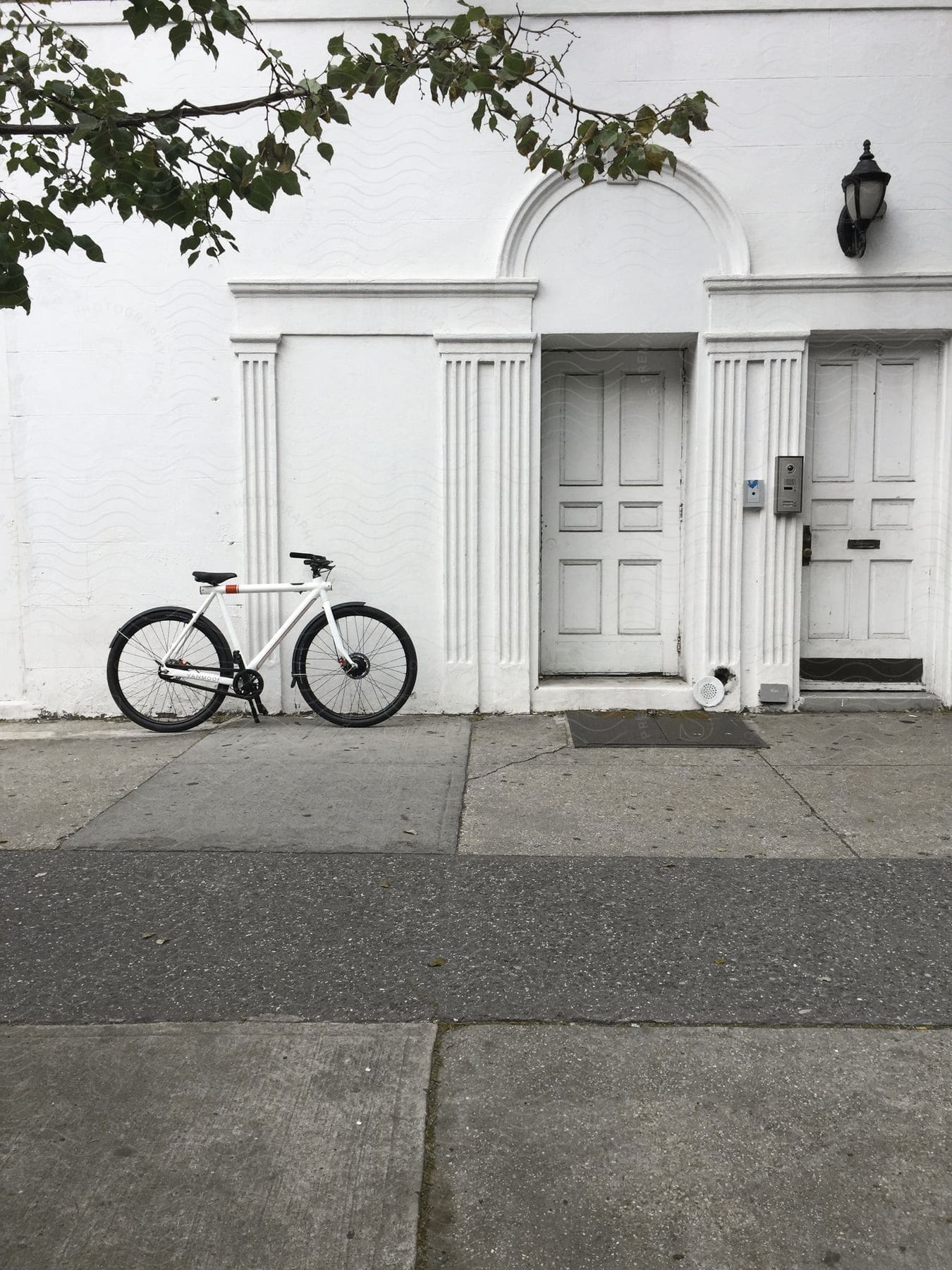 A parked bicycle next to a white building with two doors a tree branch overhead and a lamp above the door