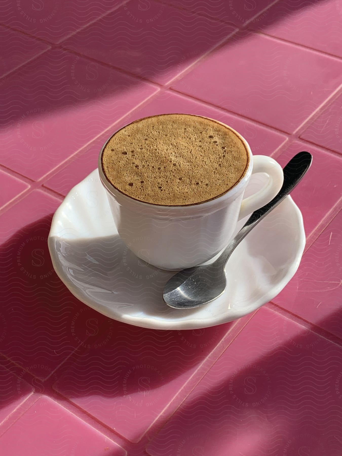 A cup of coffee sits on a pink tile floor undisturbed with its saucer and spoon