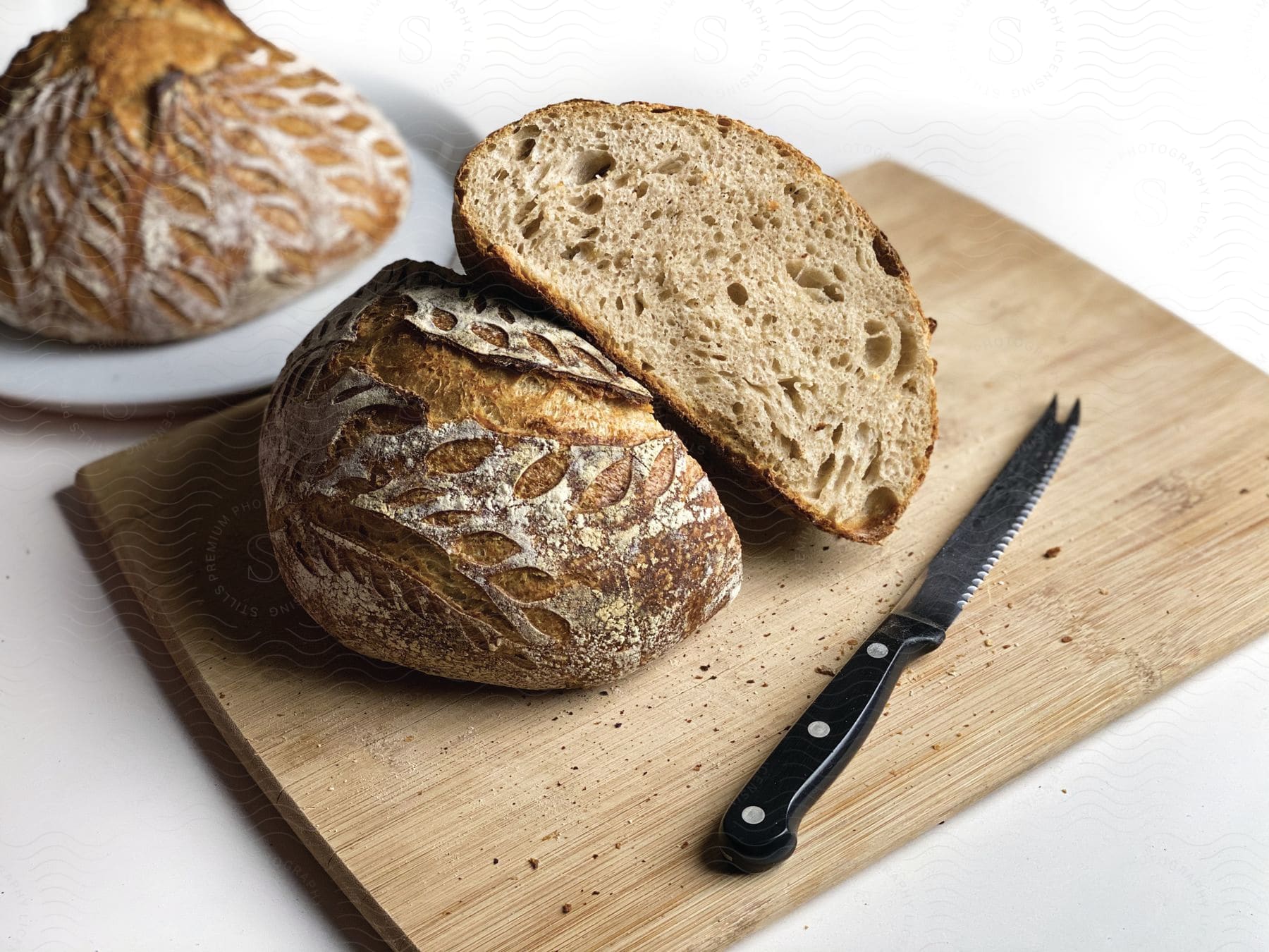 A loaf of bread cut in half on a cutting board with a knife and another loaf of bread on a plate in the background