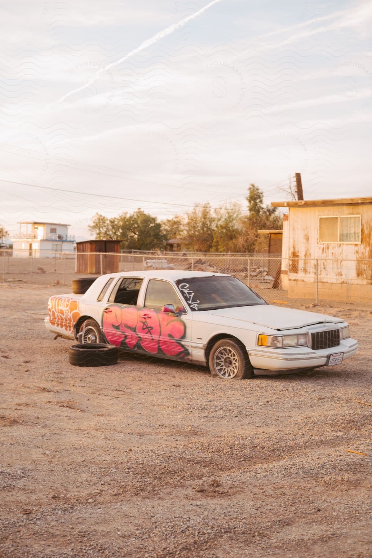 An abandoned car with broken windows and flat tires in an empty parking lot with graffiti painted on it
