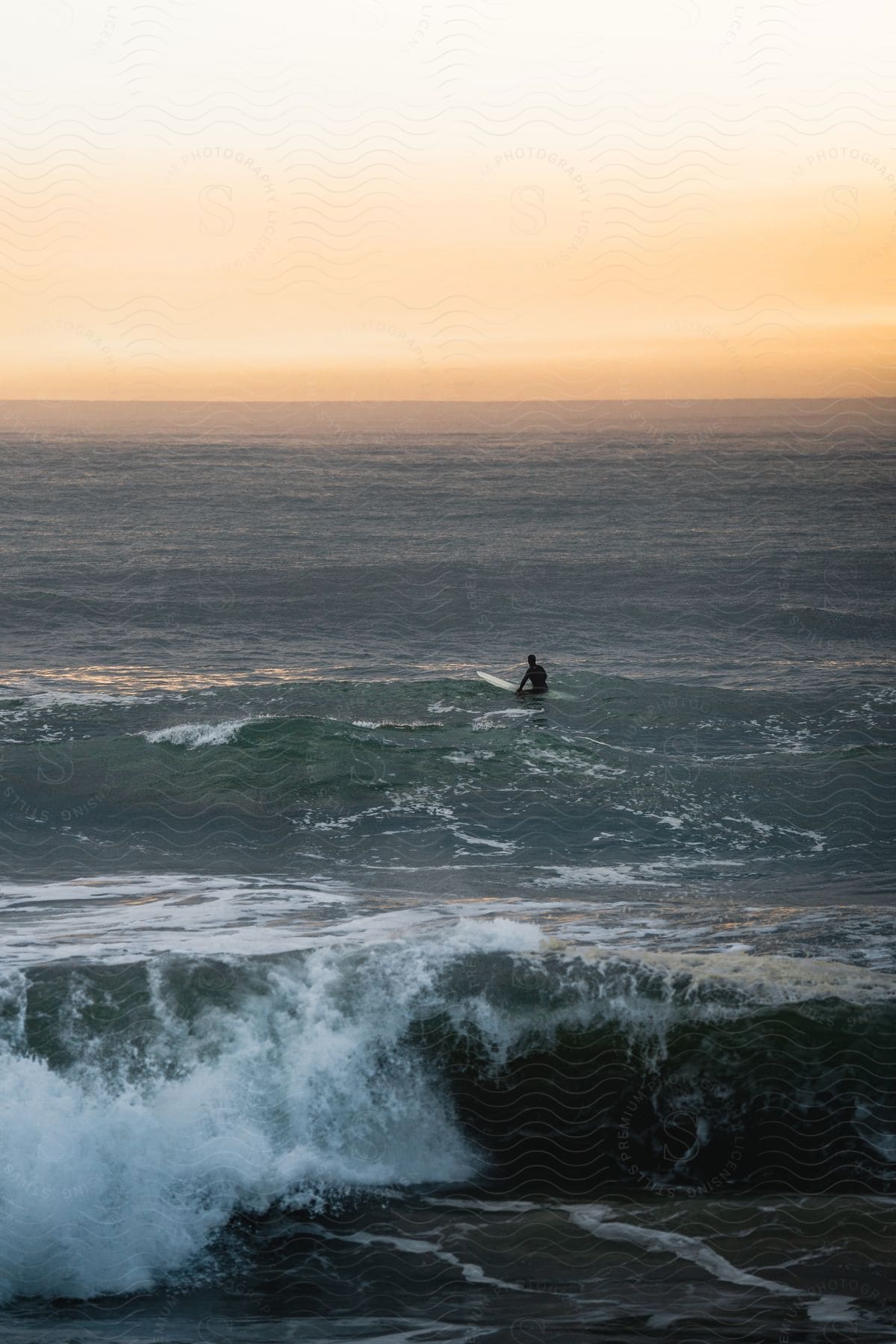 A man sits on his surfboard facing an orange glow on the horizon as waves roll into shore