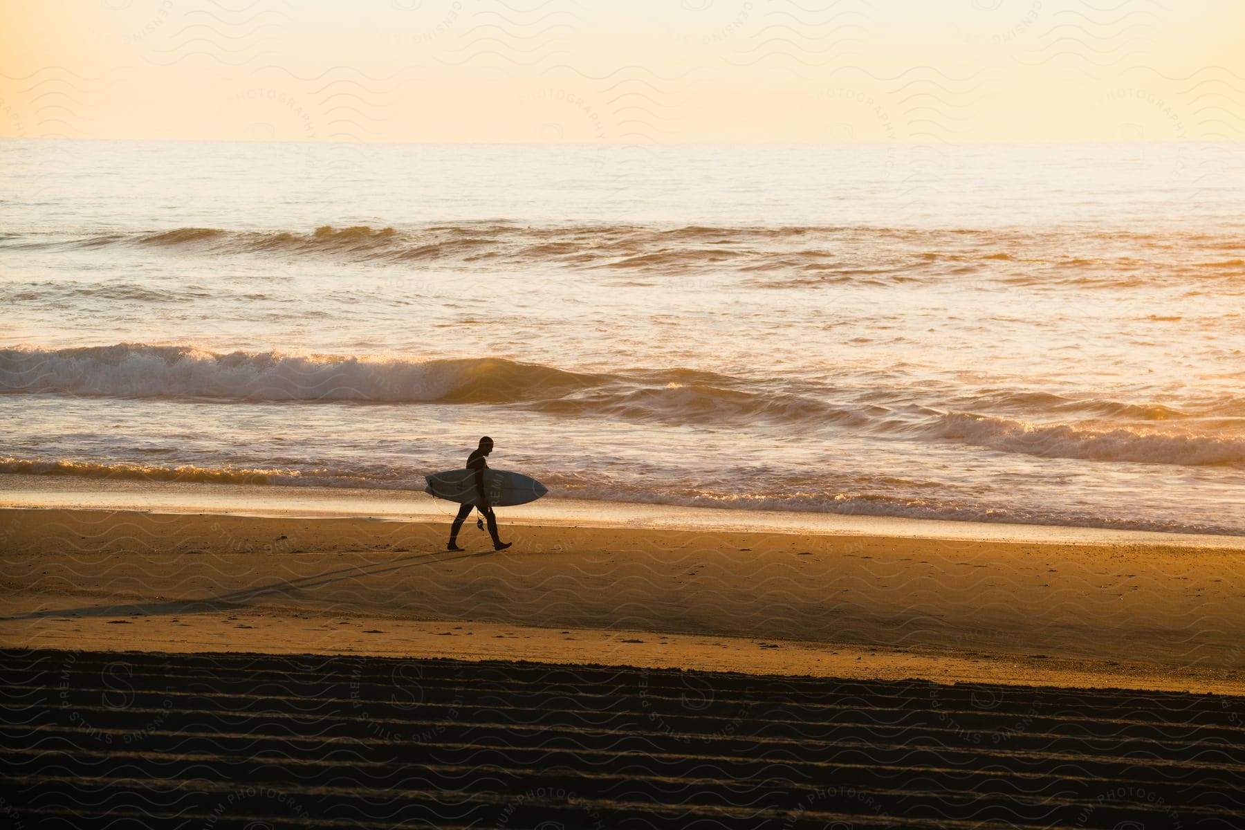 A person in their 20s or 30s wearing a wetsuit is carrying a surfboard on the beach near the ocean at sunset