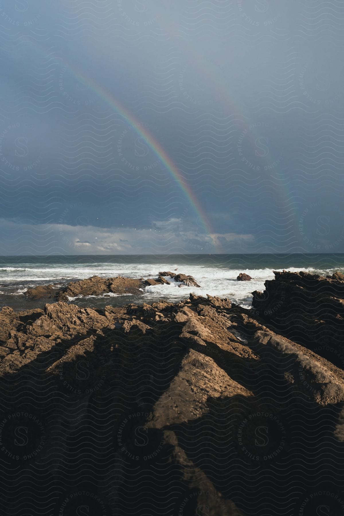 Double rainbows appearing in the cloudy sky above the ocean coastline