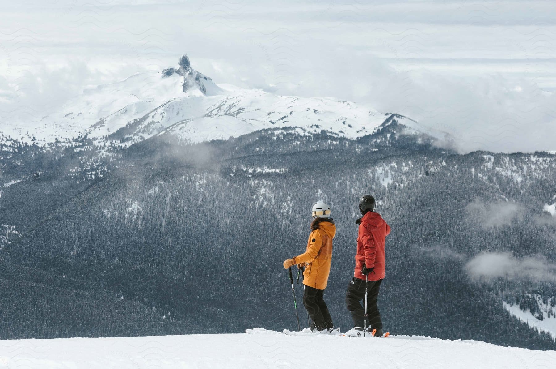 Two skiers in winter gear standing on a mountain looking towards another mountain