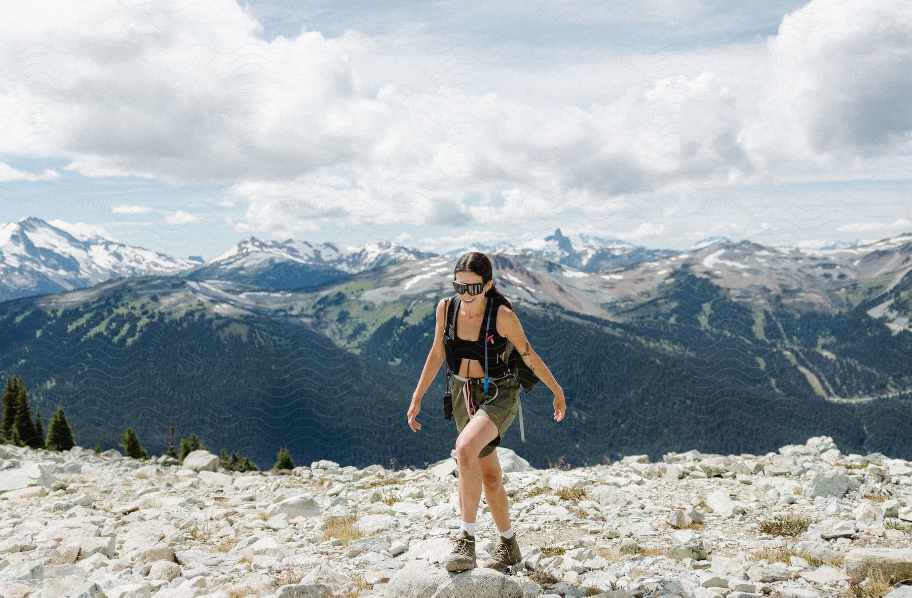 A woman walks on rocks as she hikes across a mountain with mountains in the distance under a cloudy sky