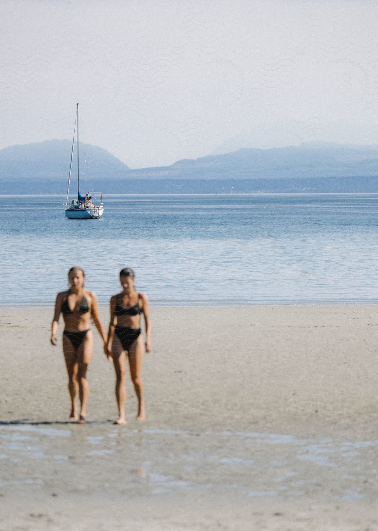 A sailboat floats off the coast while two women in bikinis walk on the beach