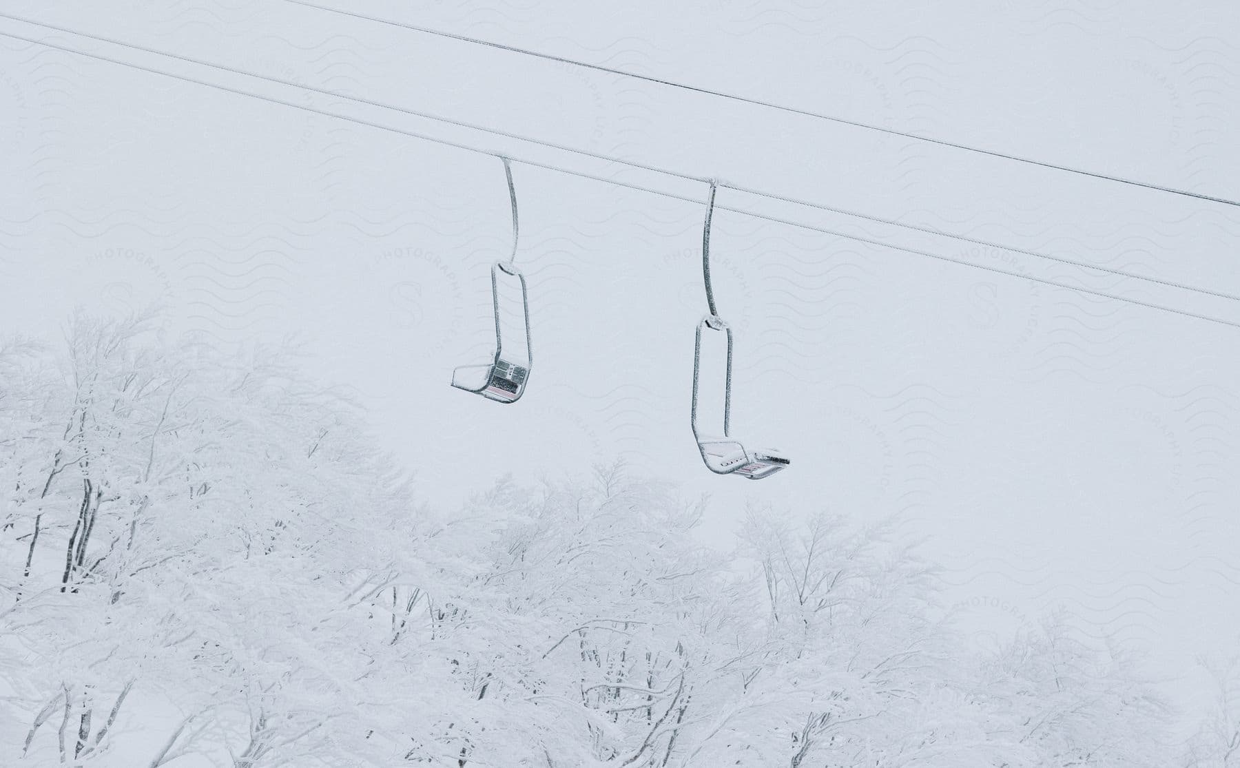 Empty ski lift seats hang from cables with snowcovered trees in the distance