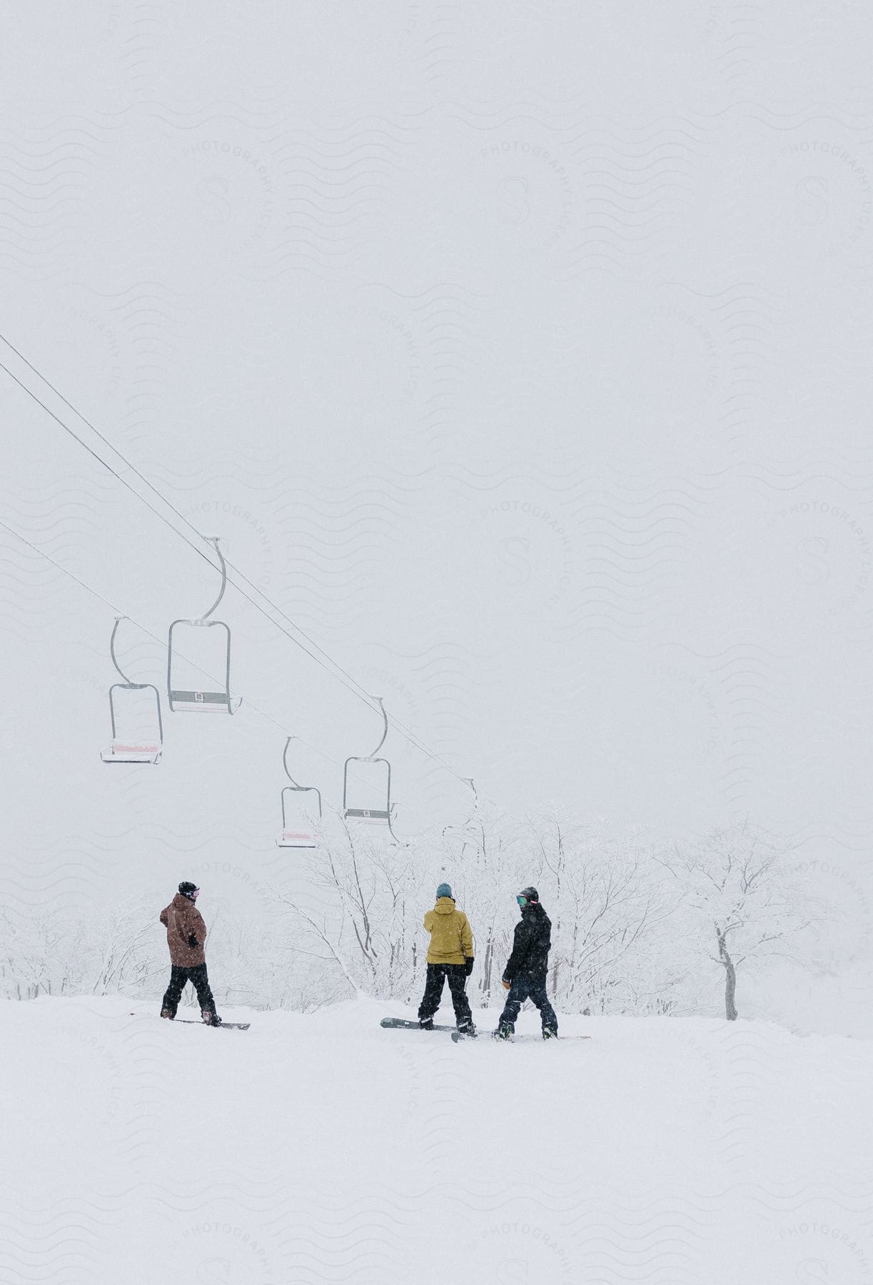 Three snowboarders stand on the snow under chairlifts under a white sky