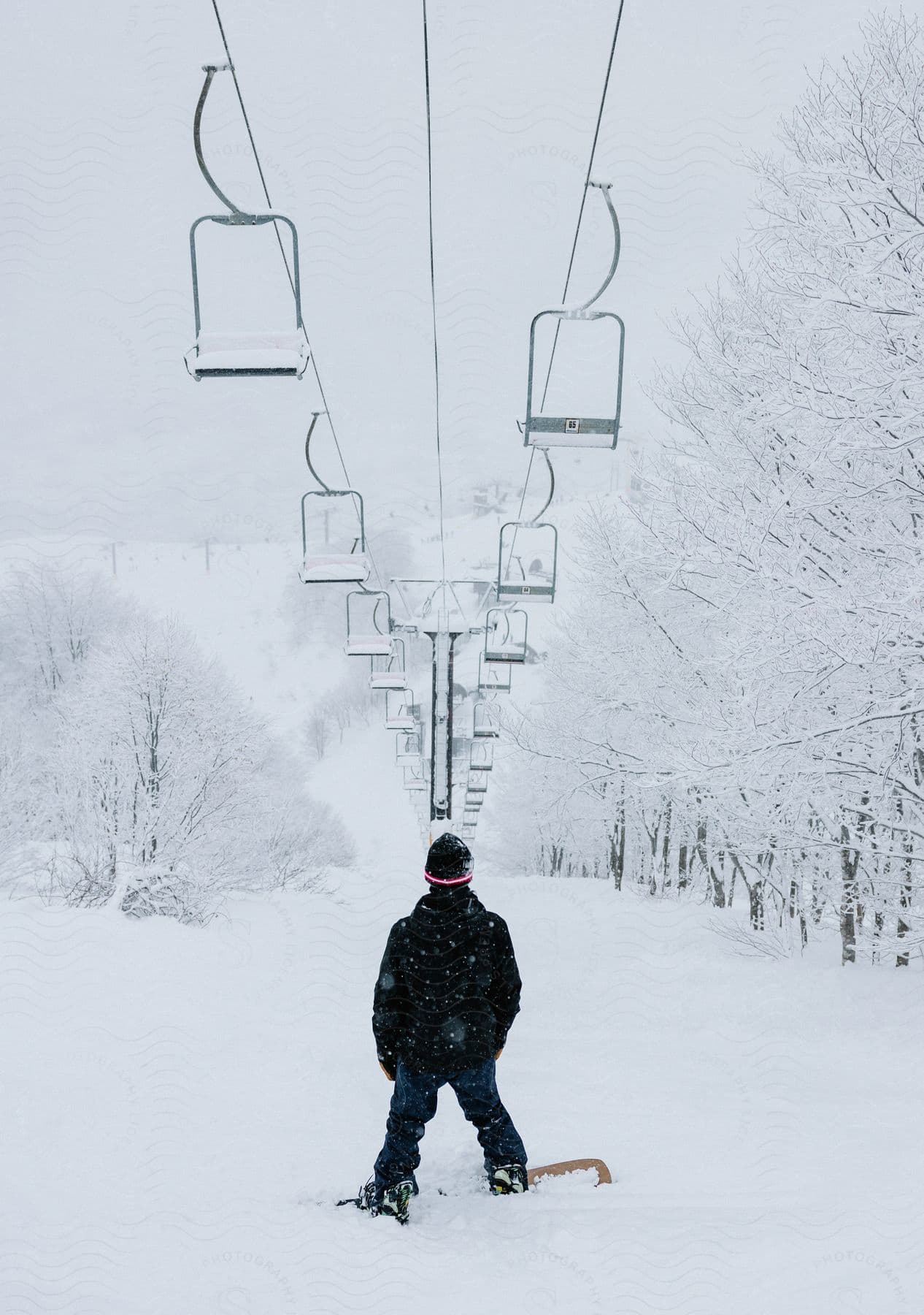 A snowboarder stands on a slope underneath a ski lift