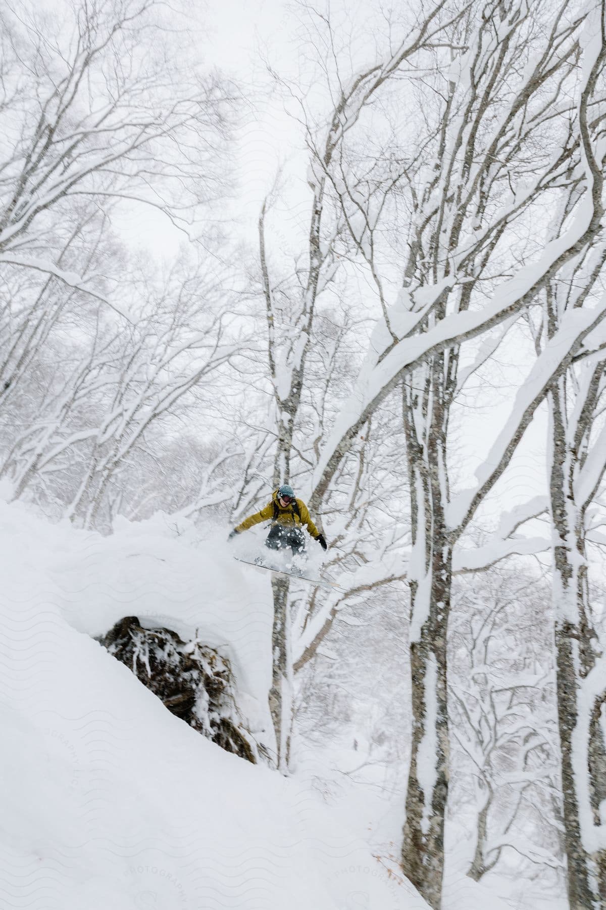 A snowboarder zooming down a snowy mountain dodging trees and doing a cool jump over a rock