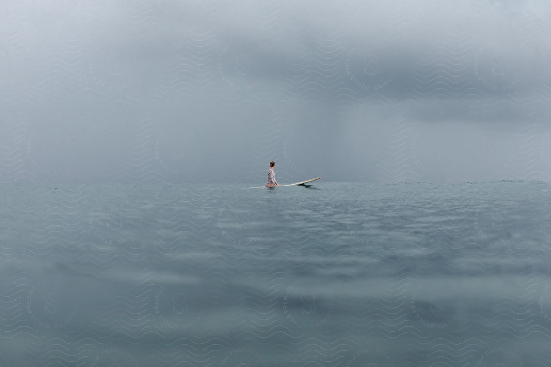 A person sits on a surfboard in the middle of the sea under a gray sky