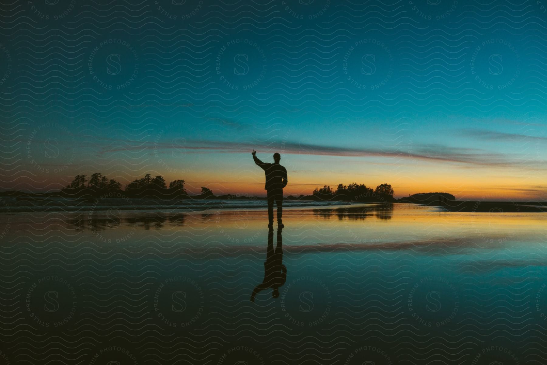 A mans silhouette and reflection on thin water with a setting sun reflecting off the water behind him and the sky getting darker to the left