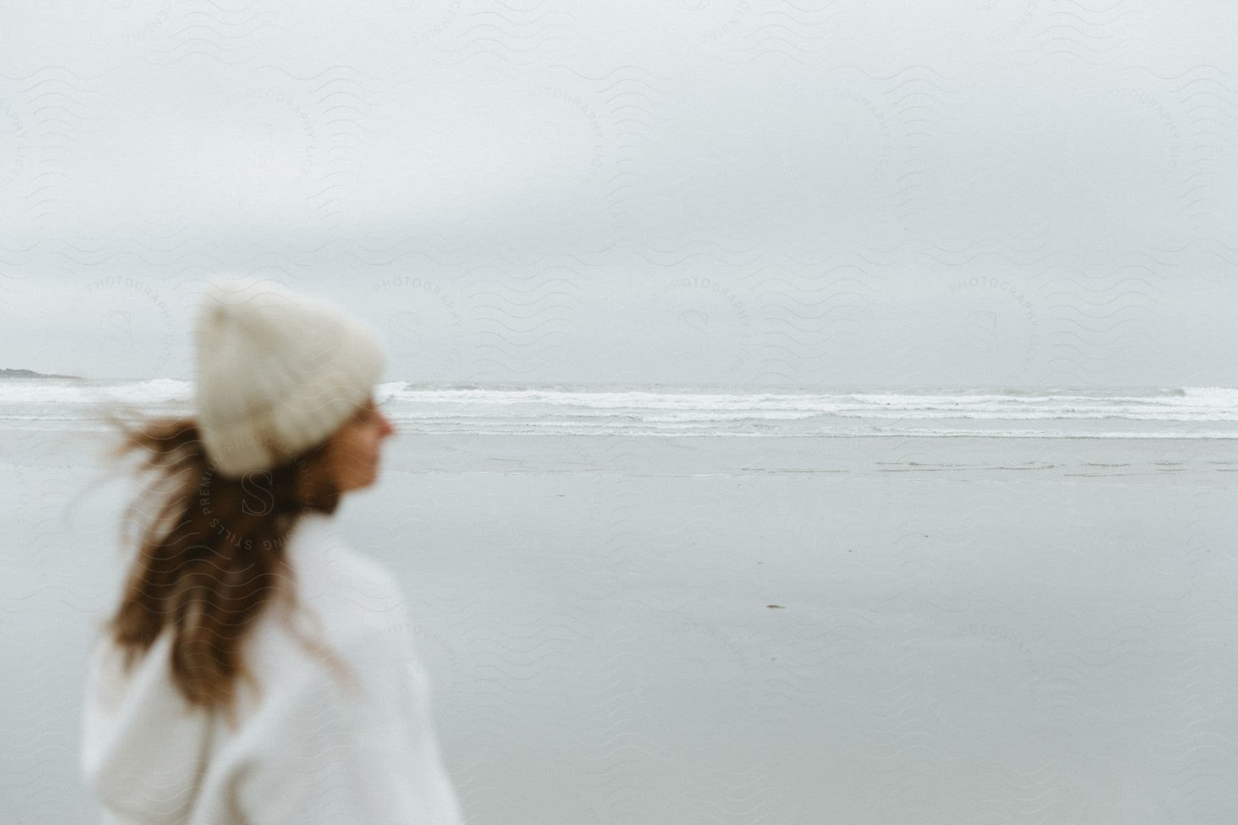 A woman wearing white warm clothes stands on a beach on an overcast day