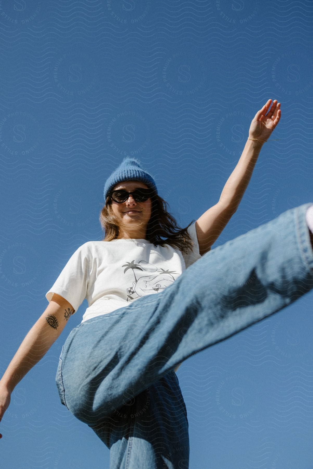The sun shines on a woman wearing sunglasses and a knit hat standing on one leg under a clear blue sky with one arm and a leg in the air