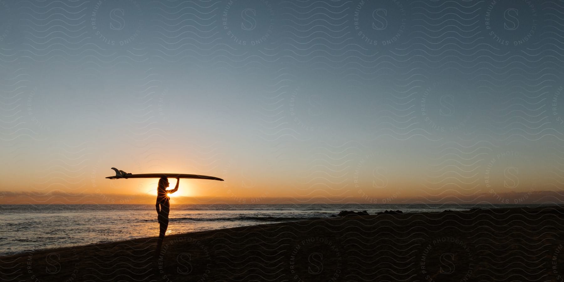 A woman on a beach holds a surfboard on her head as the sunset shines brightly in the distance