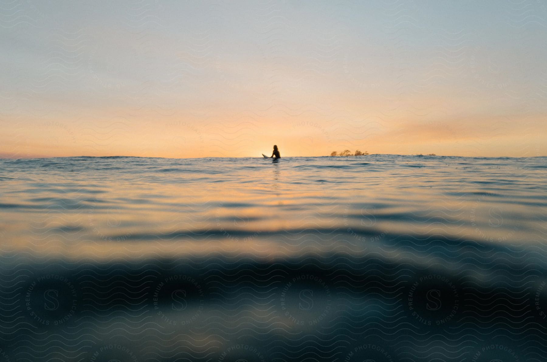 A person sitting on a surfboard on water at sunset or dawn with a boat in the distance