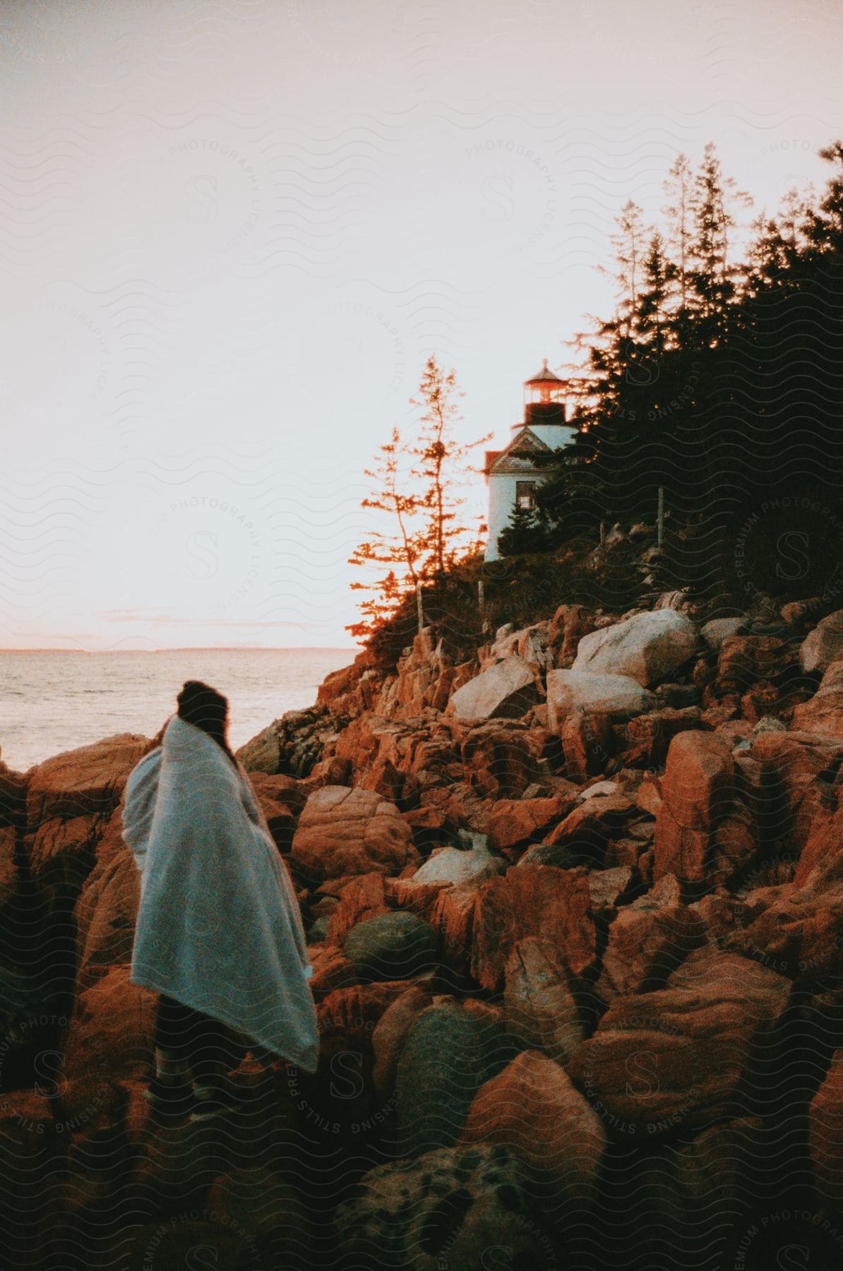 Stock photo of a woman stands on rocks near a lighthouse in maine wrapped in a blanket as the sun rises