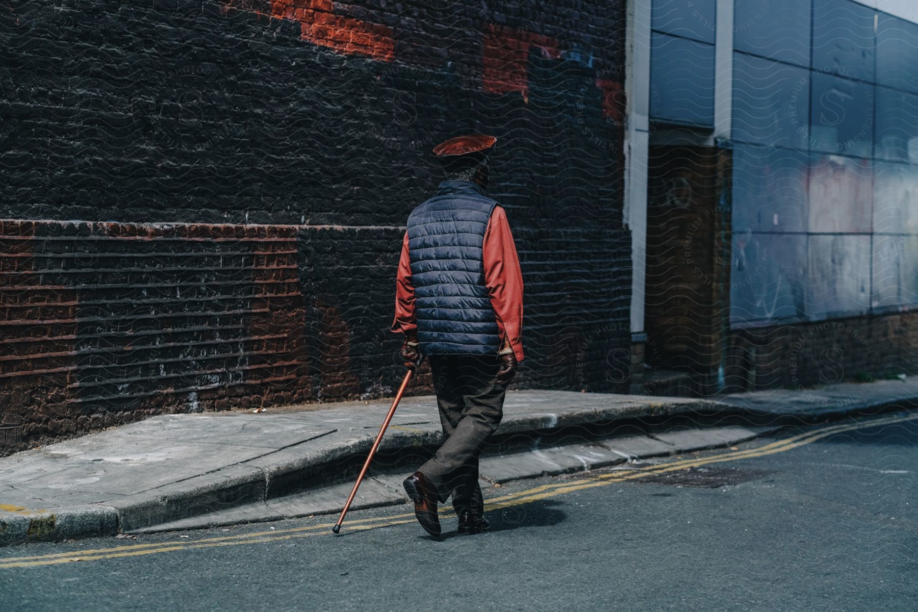 A man wearing a red shirt blue vest brown pants and a walking stick is walking down a city street