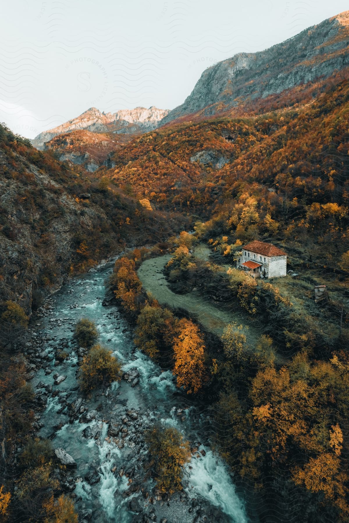 A river flows down a mountain with an older house built on the side covered with trees changing with the season