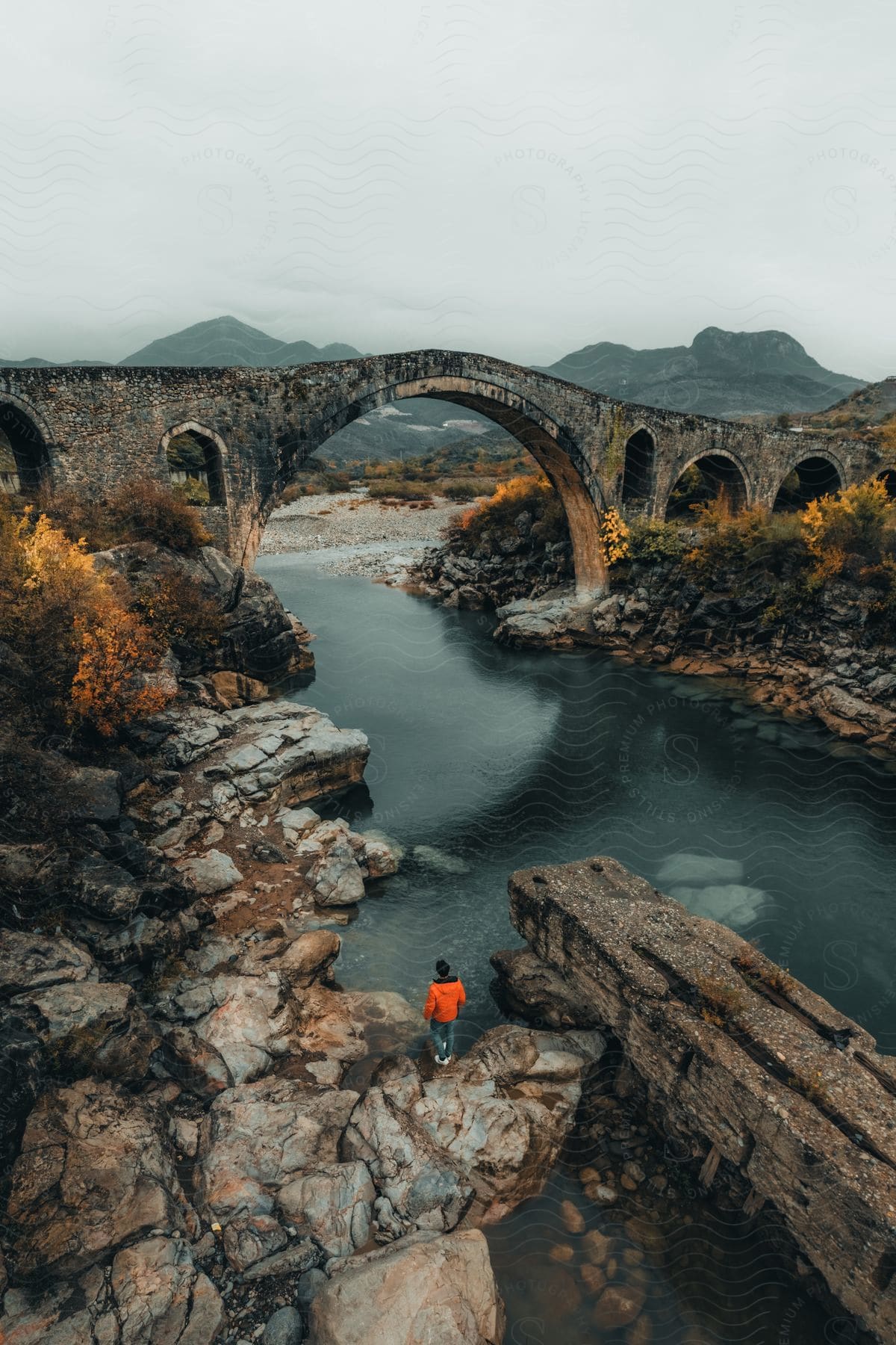 A person standing near a bridge with a lake and mountains in the background