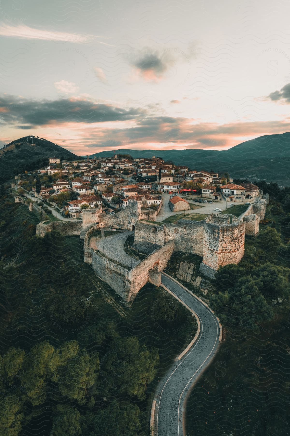 A town is situated within the walls of a stone fortress atop a forested hill near the mountains