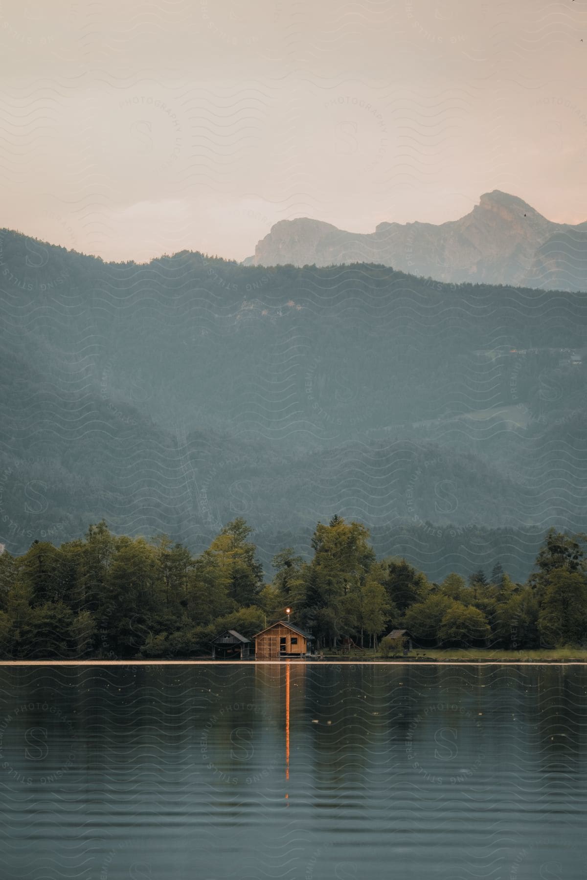 A long cabin sits on the shore of a lake surrounded by mountains and a forest tree line on a cloudy day