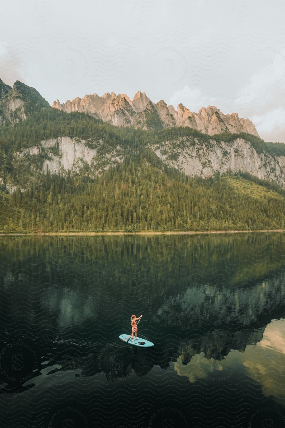 A woman stands on a paddle board on a still lake with rocky forested mountains in the background