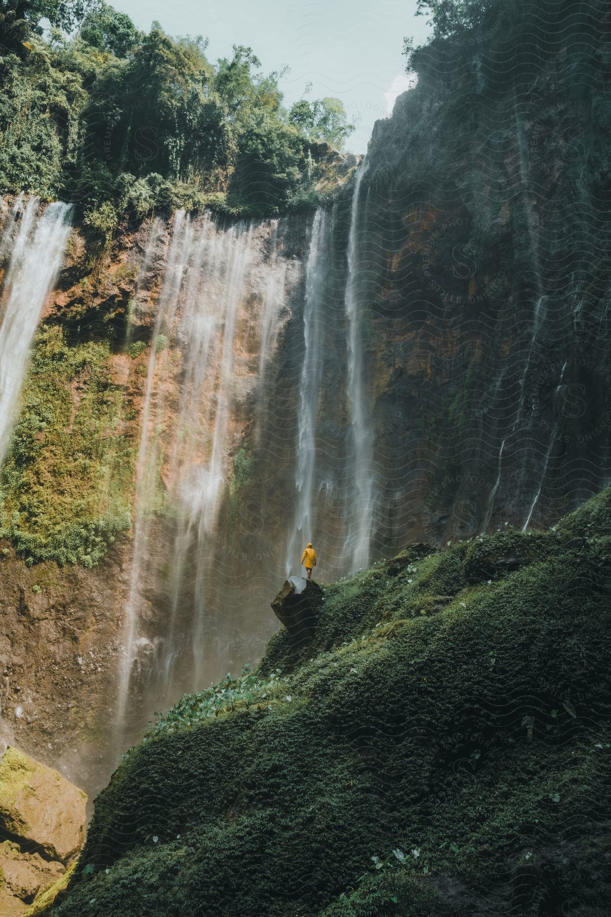 A person in a yellow raincoat walking towards a waterfall on the mountains