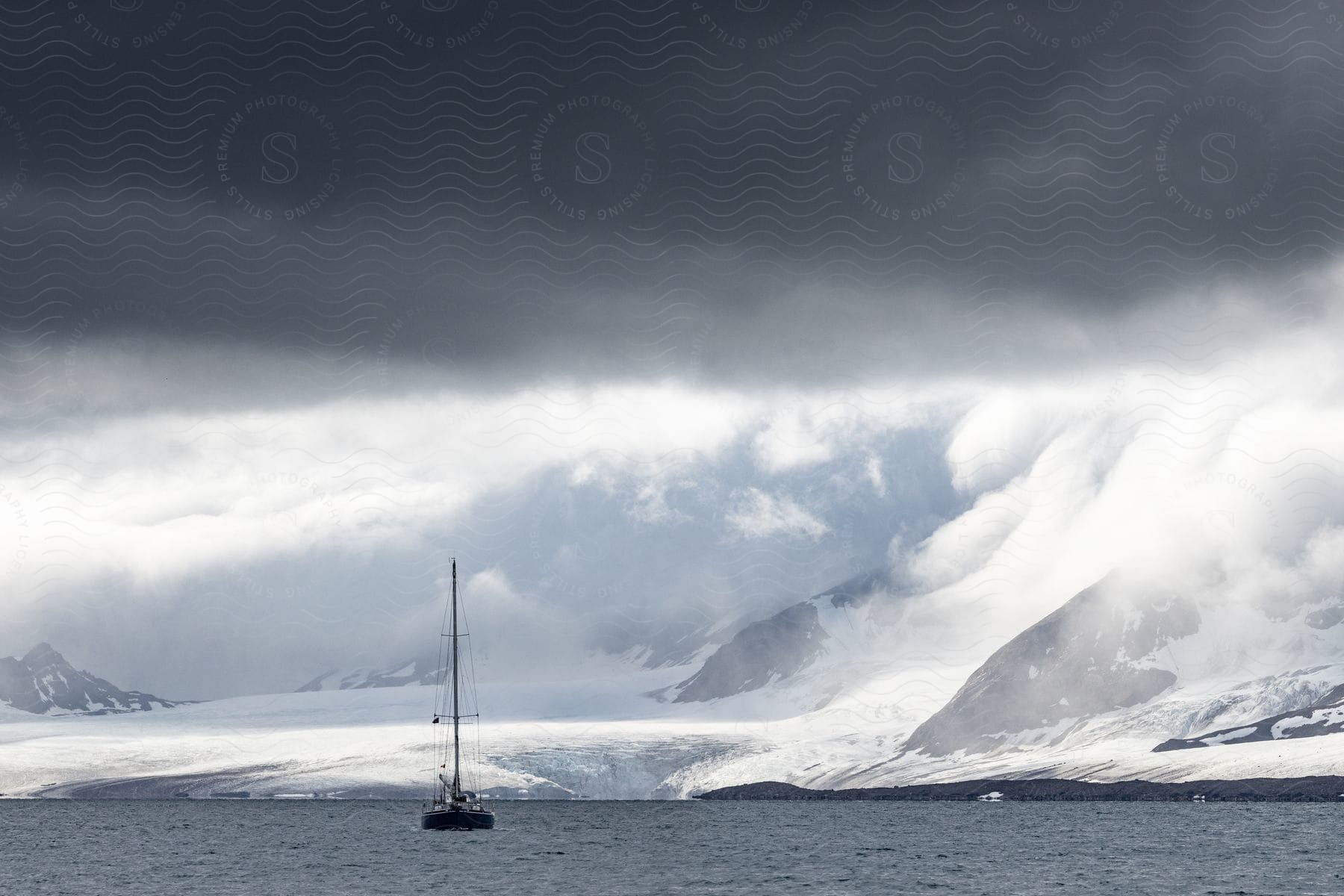 A sailboat on the water in front of a snowcovered mountain with clouds overhead