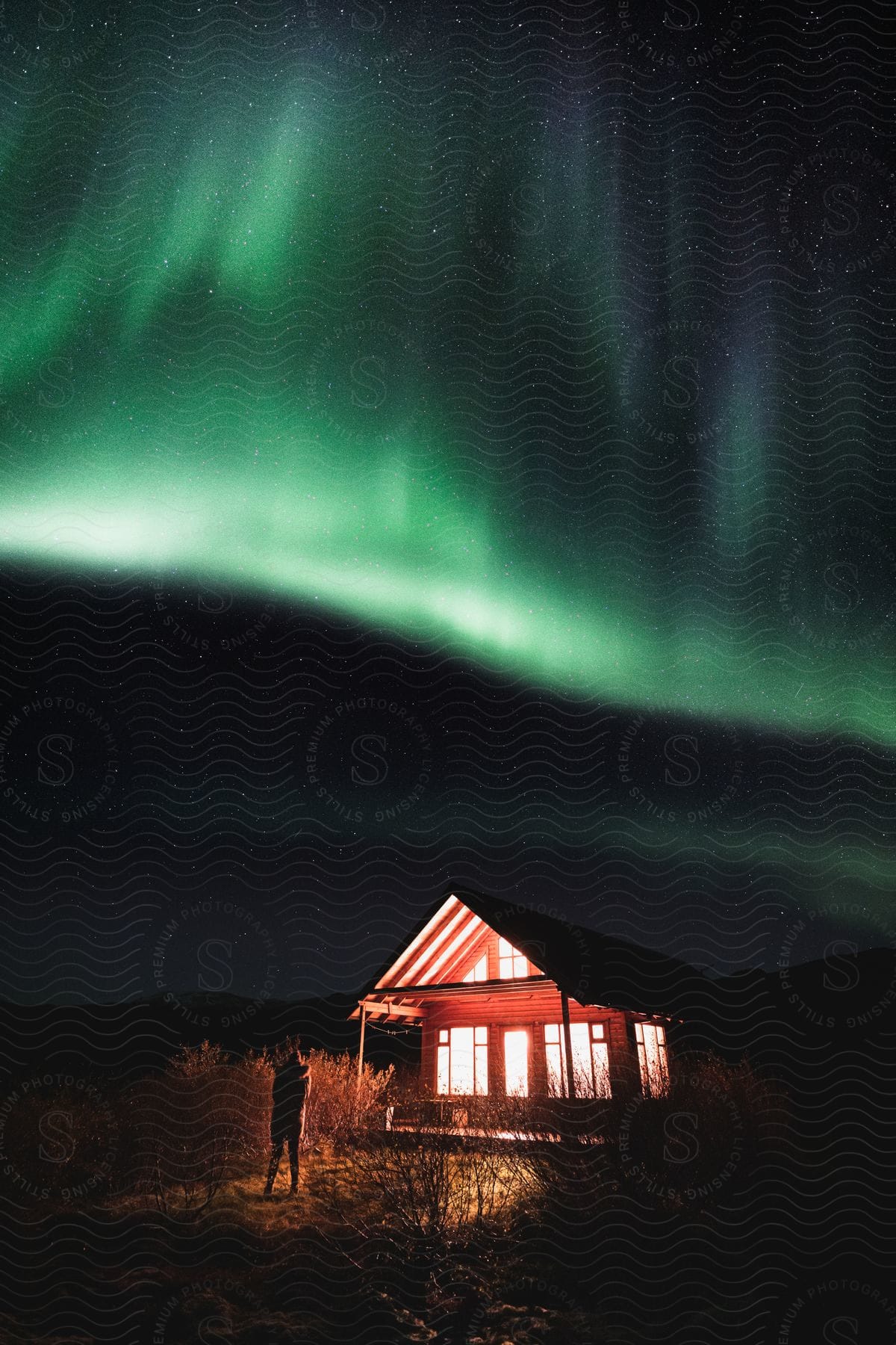 A person outside their litup home looks up at the green aurora borealis in the sky