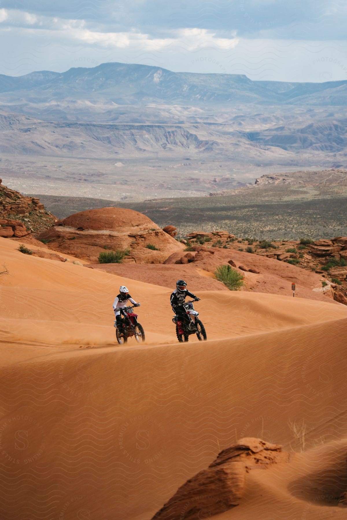 Two people riding motorbikes on a desert dune with a mountain range in the background