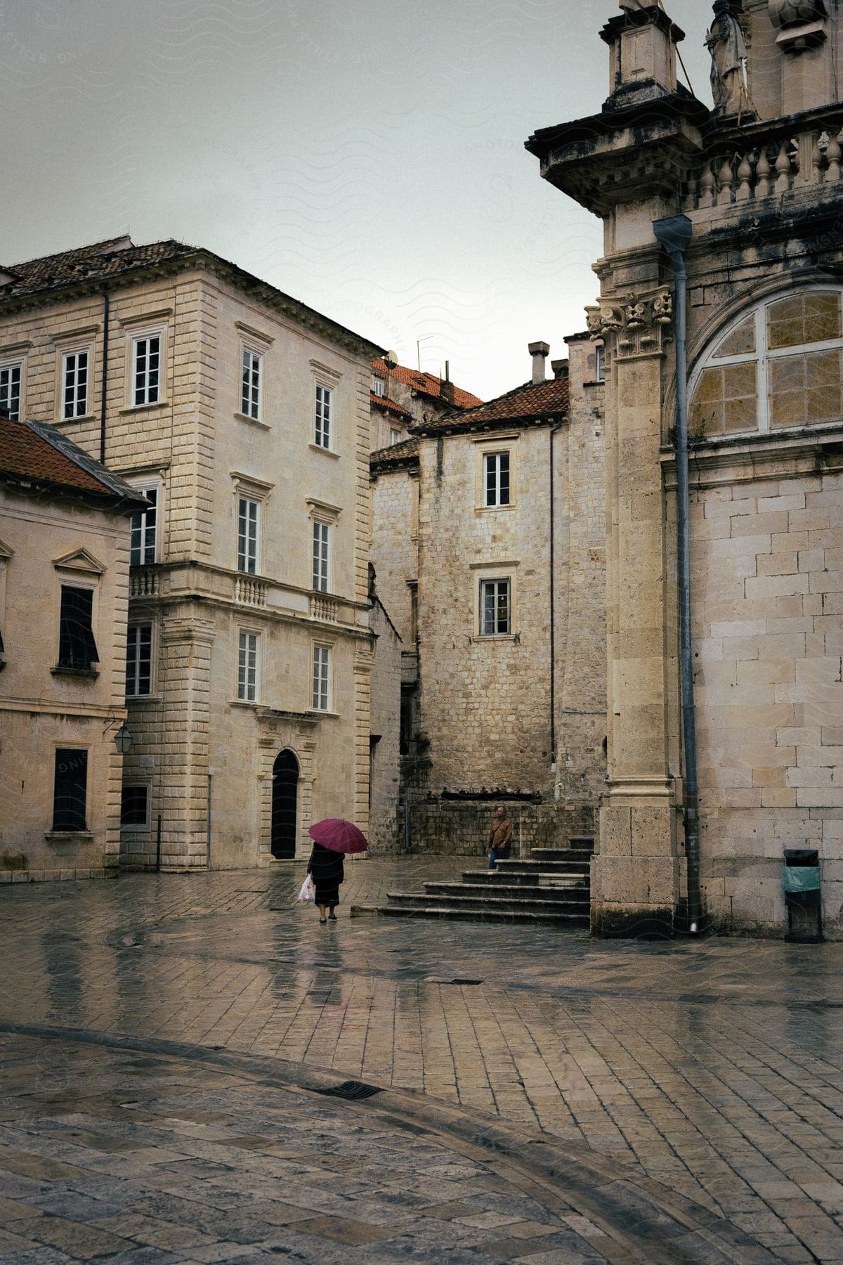 A woman walking with an umbrella near tall stone buildings and stone steps