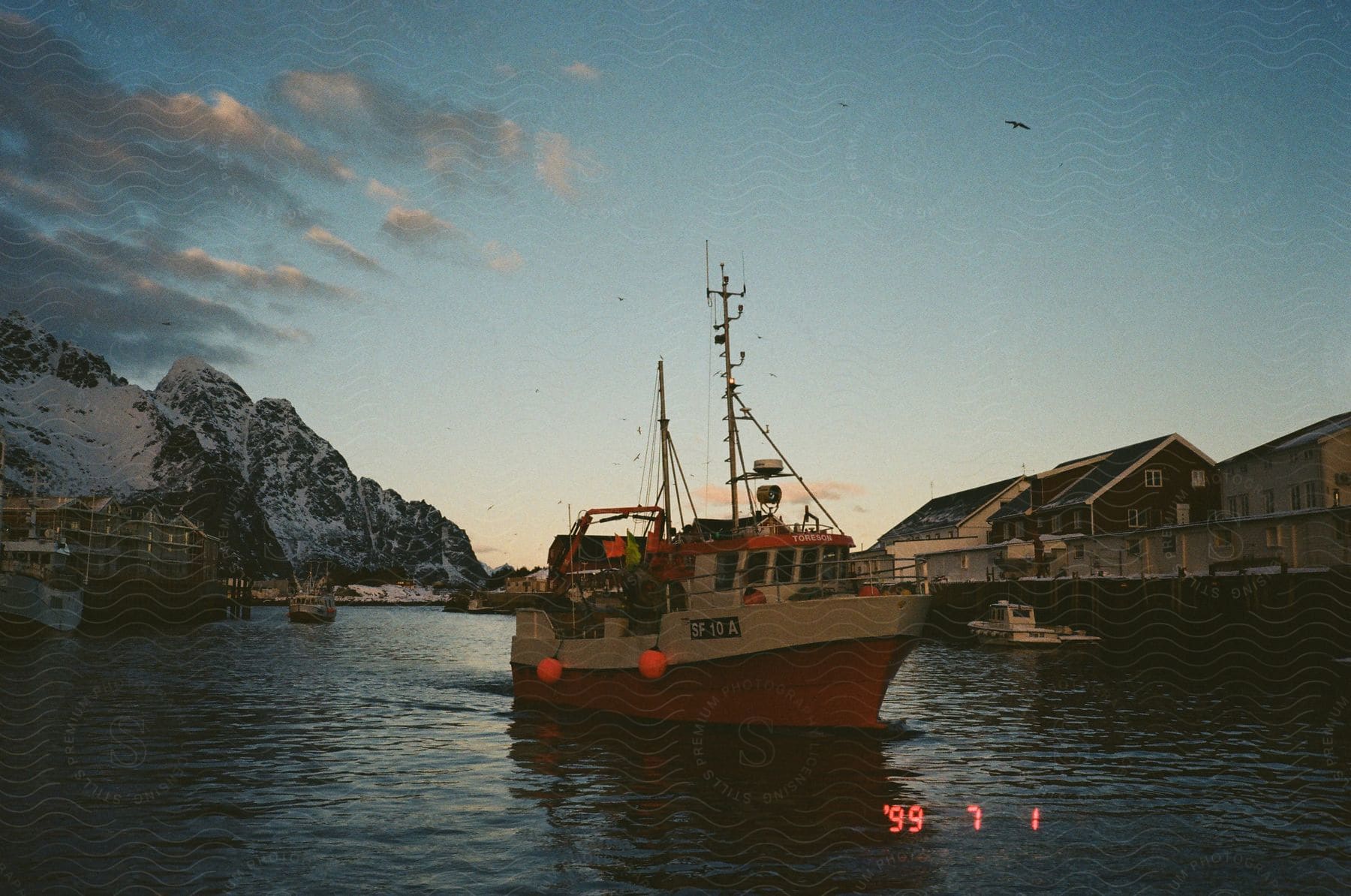 Fishing vessel floats off coast with snowy mountains and waterfront buildings at dusk