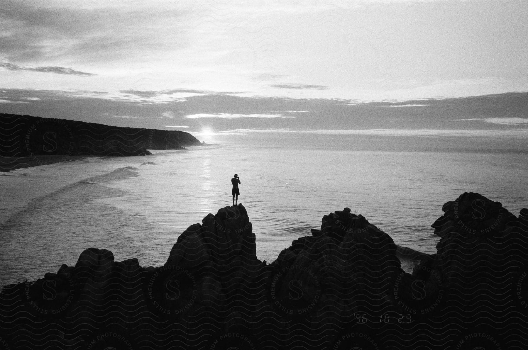 A man is standing on a rock taking a photo with a camera silhouetted against the sea and sunset