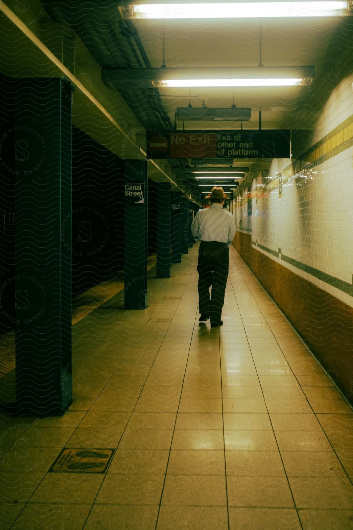 A man wearing a brown hat white shirt and black pants walks along a subway platform