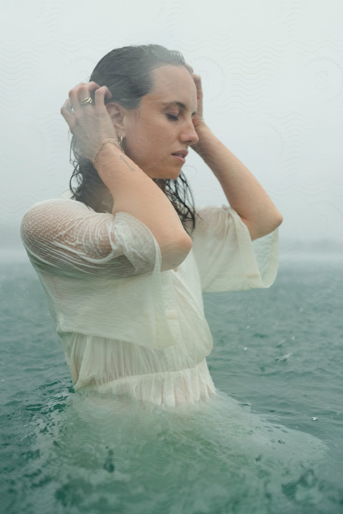 A woman in a white dress caresses her hair while bathing in a lake on a cloudy day