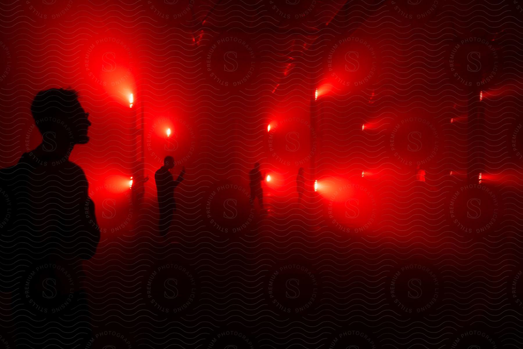 A small group of people are standing in a dimly lit warehouse with red light coming through the windows