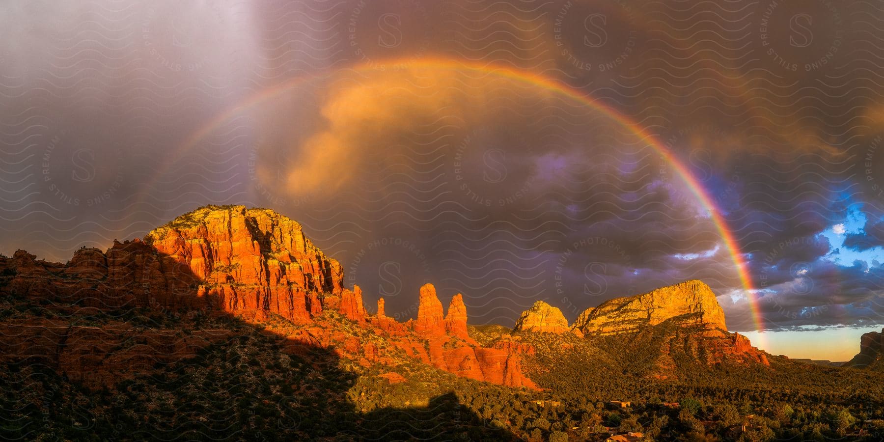 A full rainbow appears over a canyon at sunset with the shadow of the chapel of the holy cross visible at the bottom of the photo