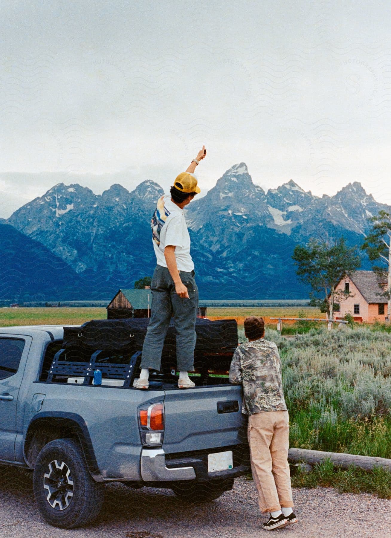 Two men standing near a truck one on the truck and the other looking at the mountain