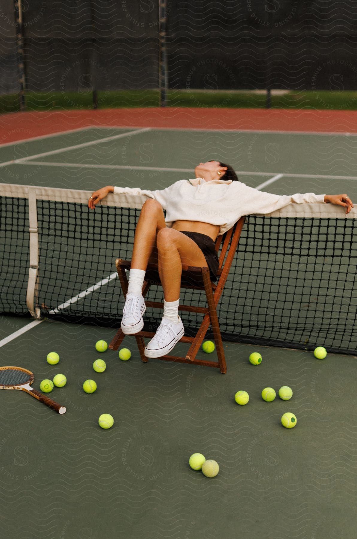 A tennis player sitting on a chair leans against a tennis net with tennis balls and rackets lying around