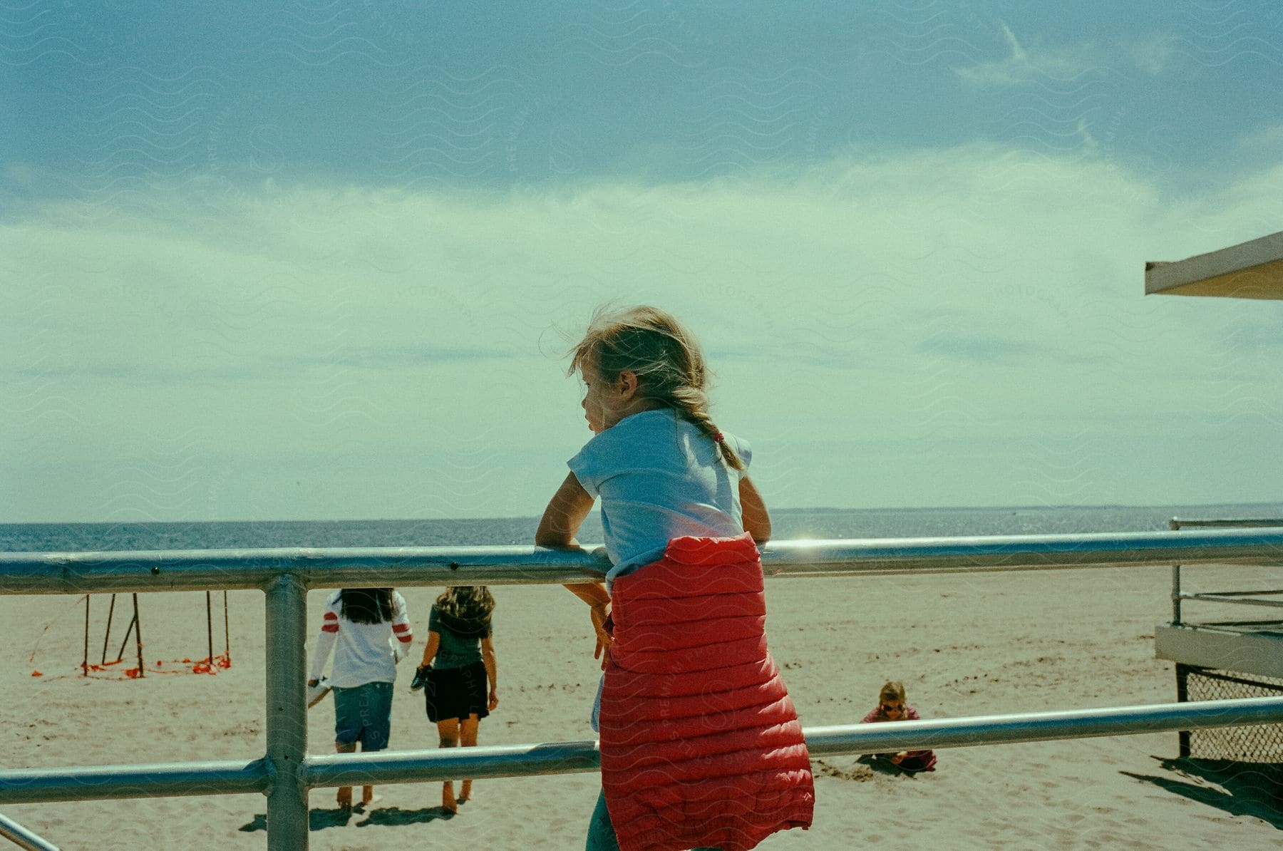 A girl leans over a beach railing watching people walk on the sand