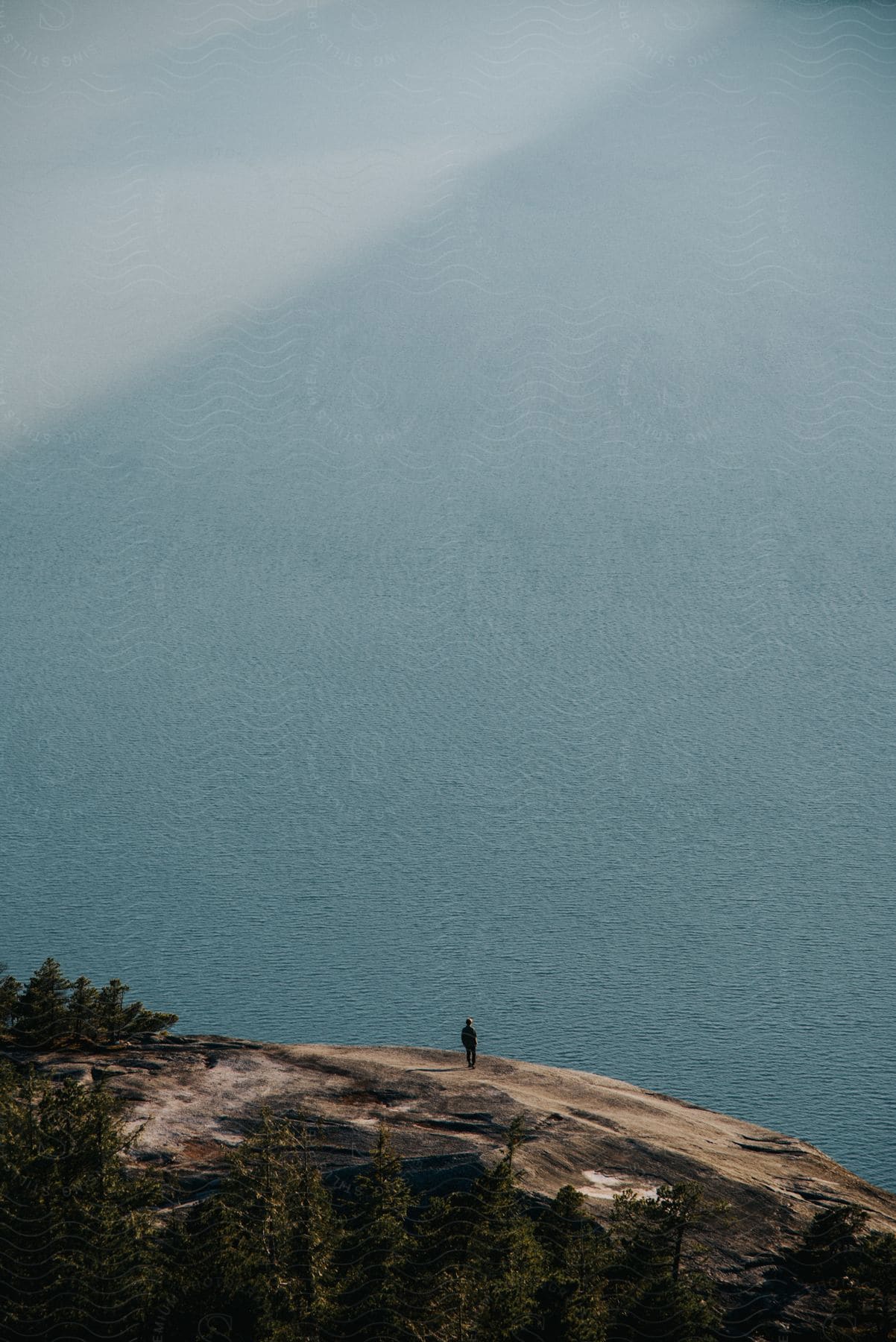 A man stands on a rocky mountain cliff overlooking the ocean under a grey sky