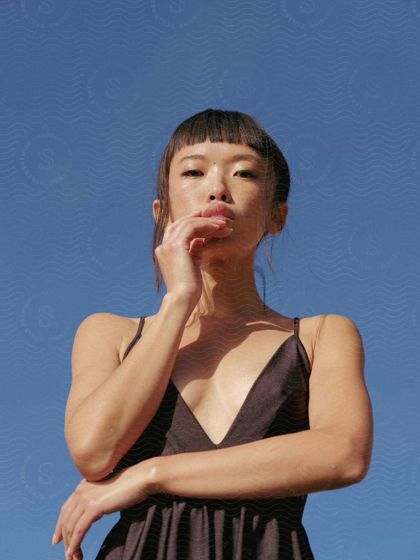 An asian model poses with her hand to her mouth wearing a brown dress and pulledback hair