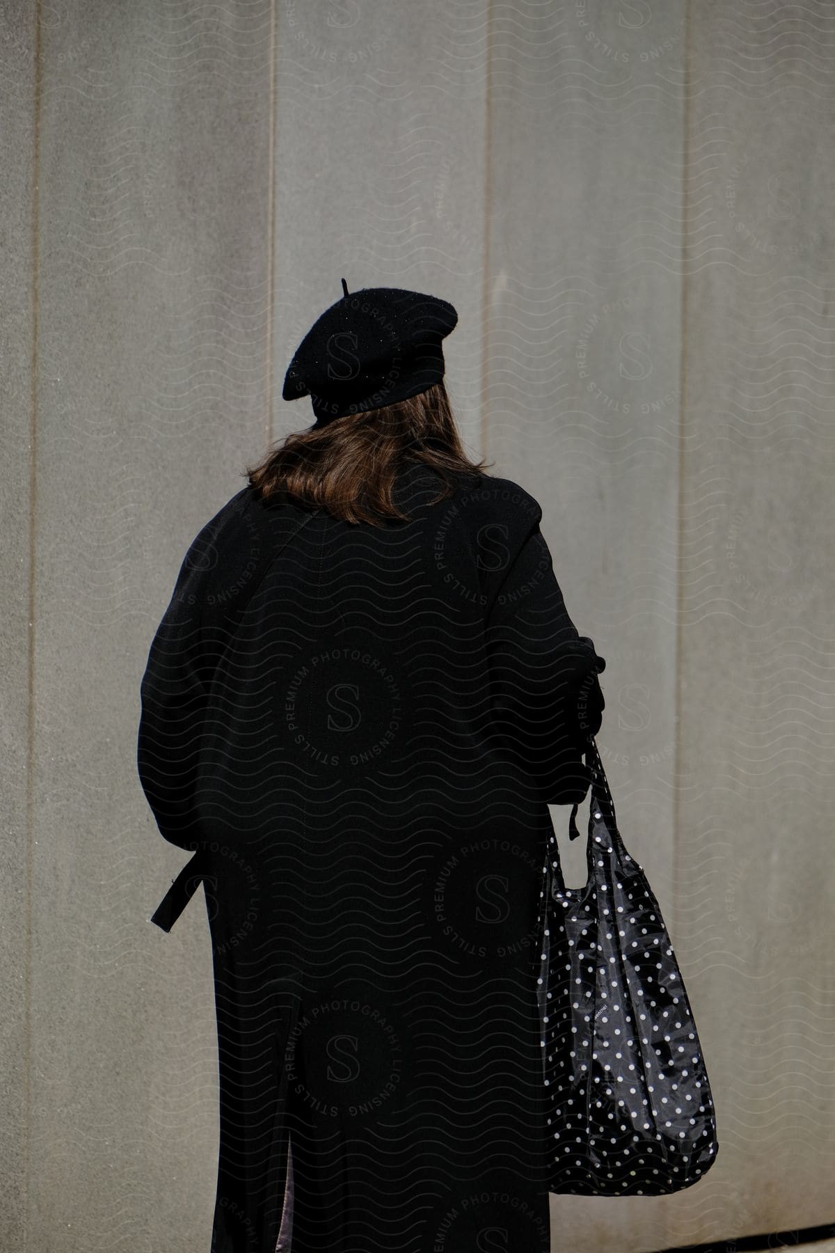 A woman wearing a long black coat a hat and a handbag walking next to a grey wall