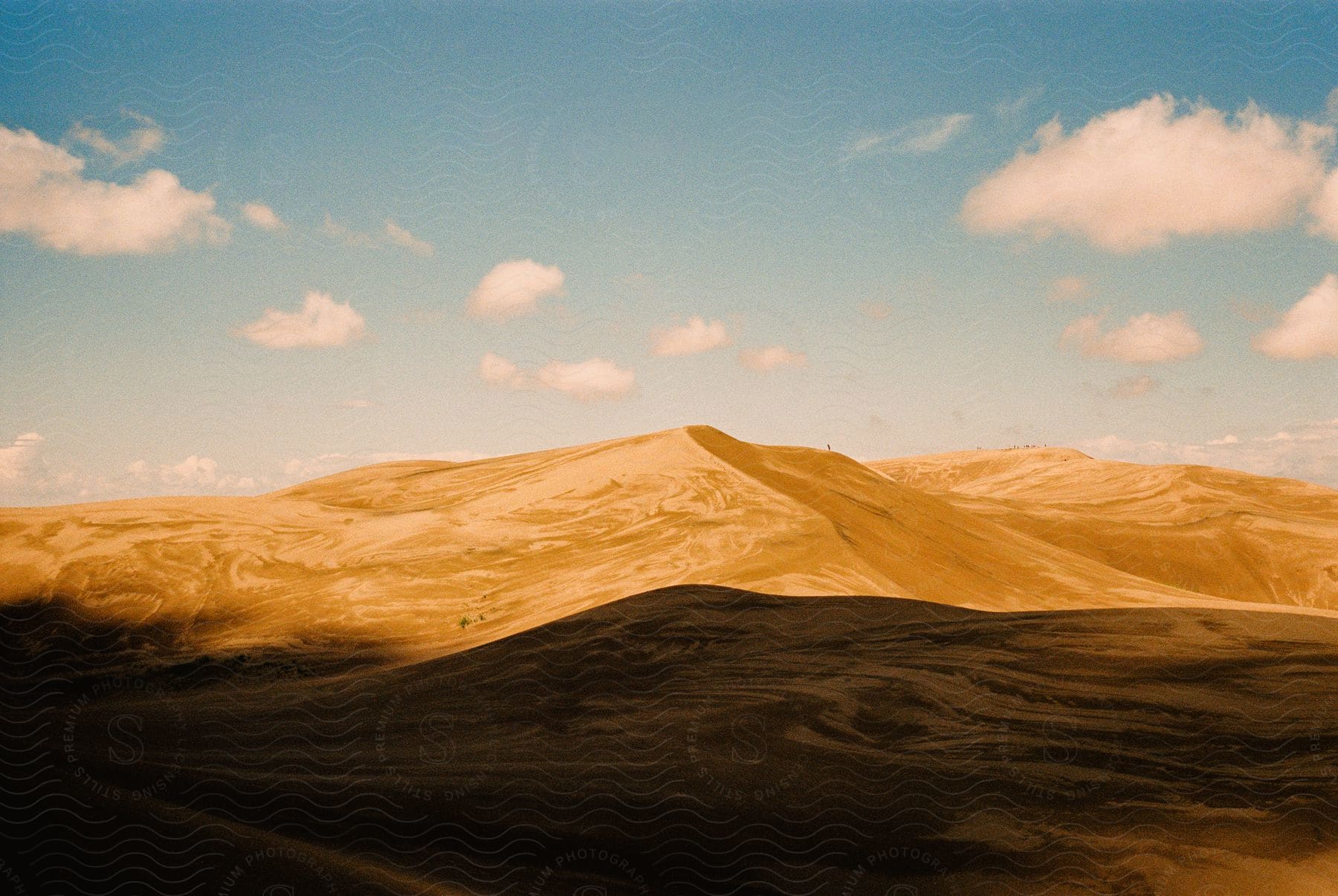 Bright rolling sand dunes with contrasting shadows against a bright blue sky with small white clouds