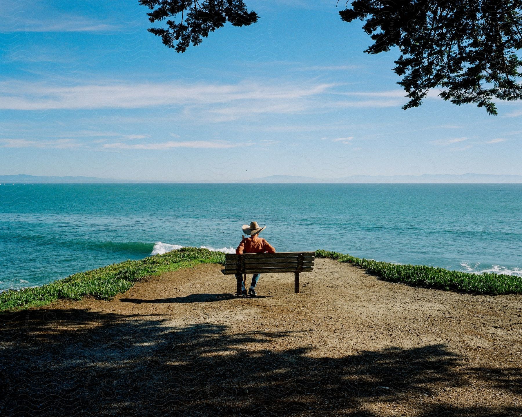A person in jeans and a hat sits on a bench overlooking an ocean on a cloudy day